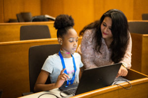 An instructor leans over a black girl's shoulder, pointing something out to her on her laptop