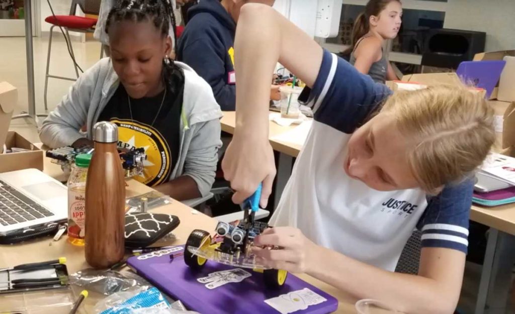 Two teen girls seated side-by-side use tools to build their robotics