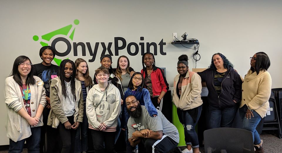 A group of girls and instructors smiling for the camera in front of a display wall that reads Onyx Point